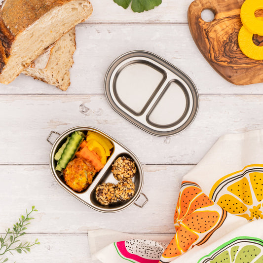 Stainless Steel lunch container with compartments filled with food on a wooden surface with bread and a colourful towel.