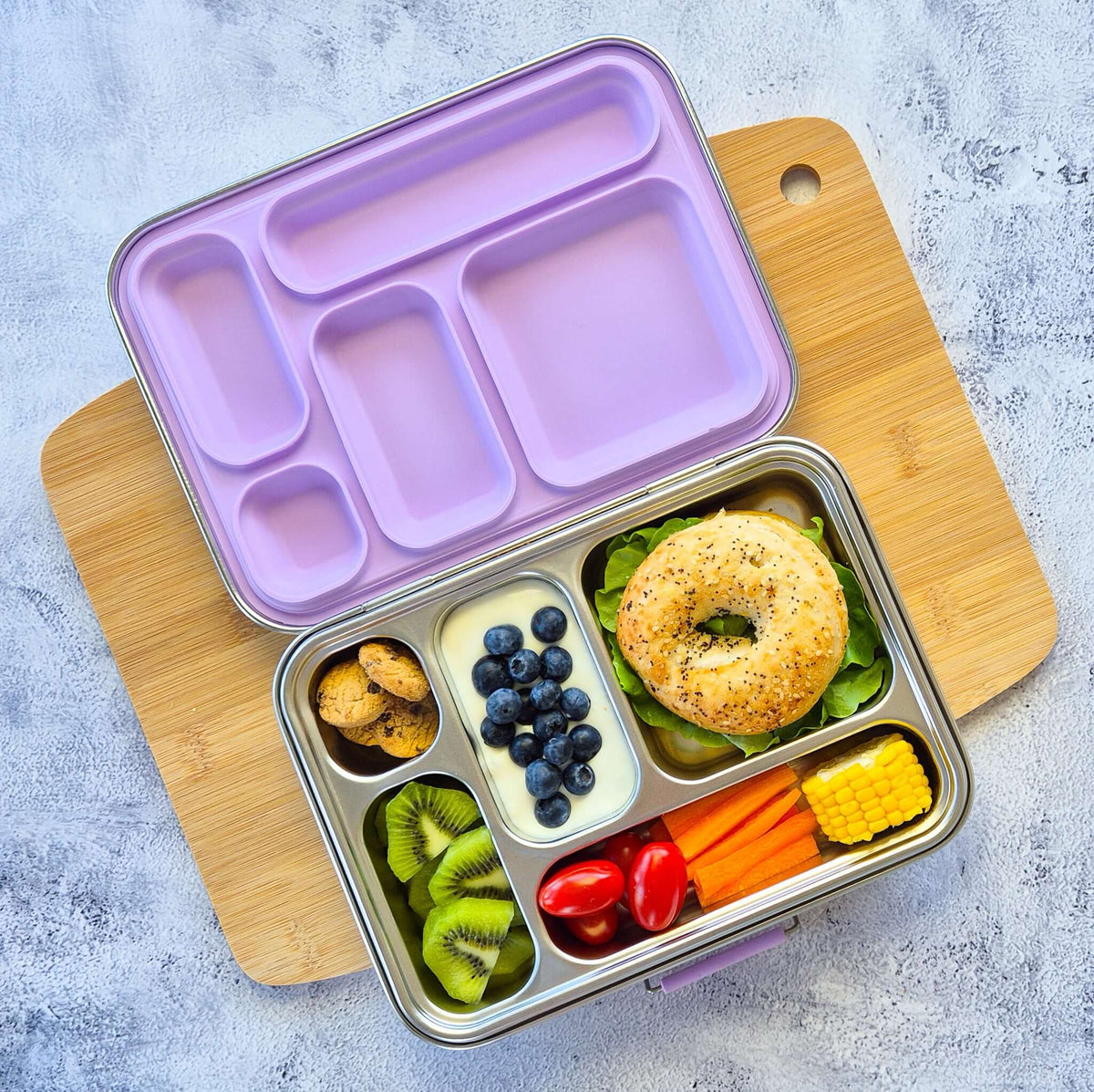 Five-compartment bento box with a lilac lid on a wooden board, packed with a bagel, blueberries, kiwi and corn