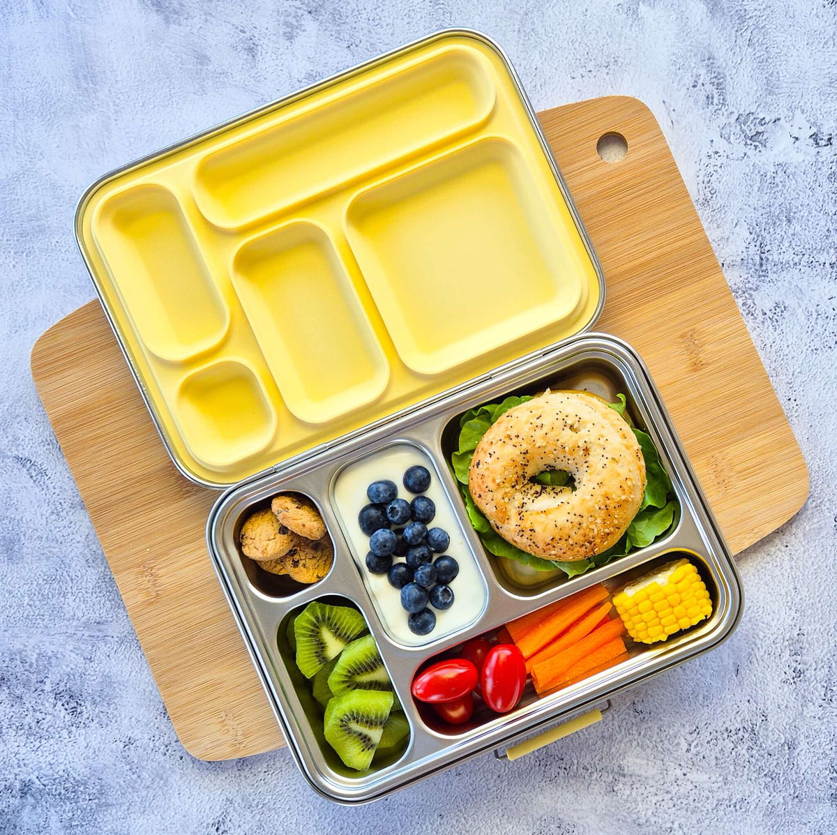 Five-compartment bento box with a yellow lid on a wooden board, packed with a bagel, blueberries, kiwi and corn