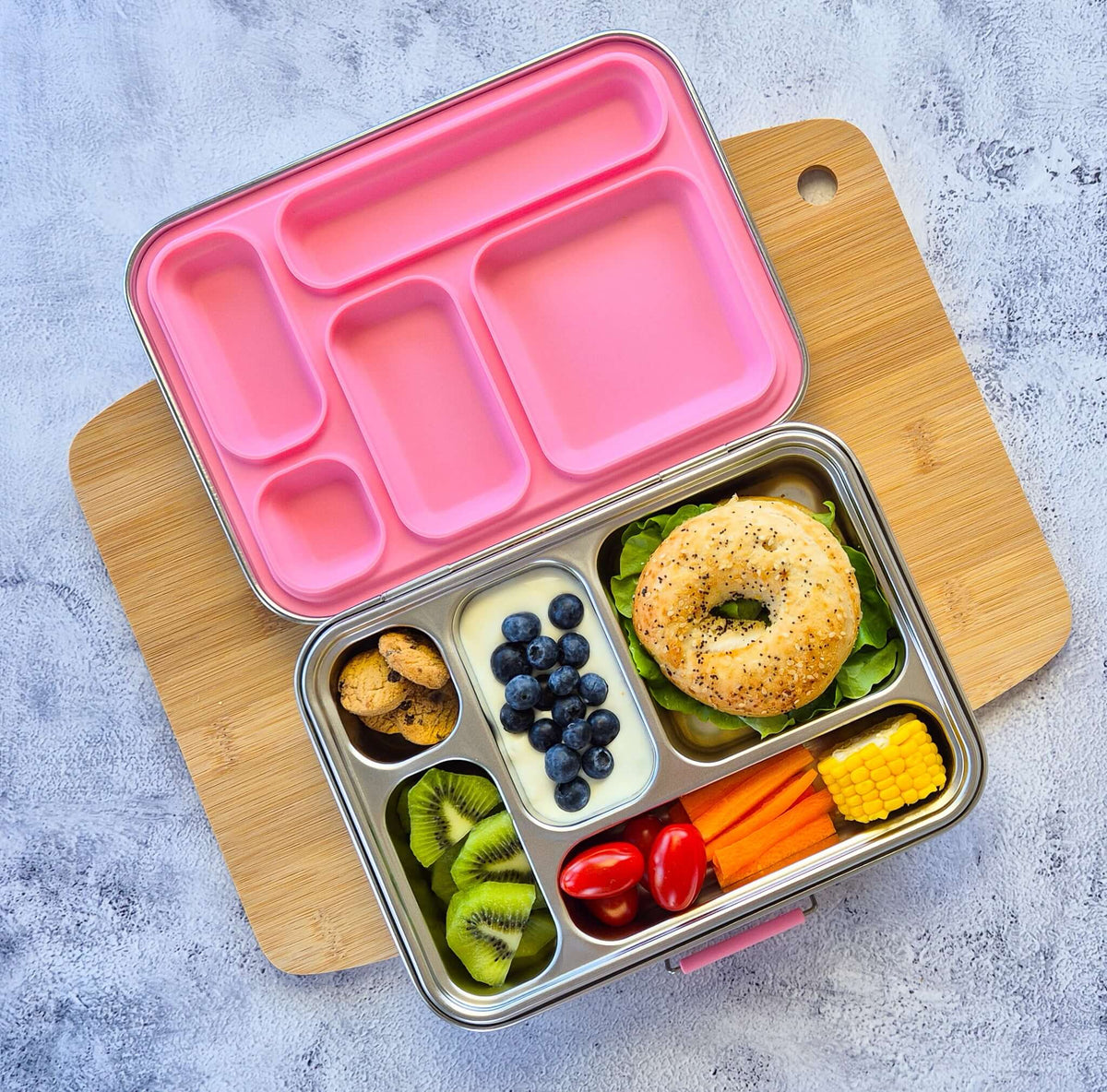 Five-compartment bento box with a pink lid on a wooden board, packed with a bagel, blueberries, kiwi and carrot sticks