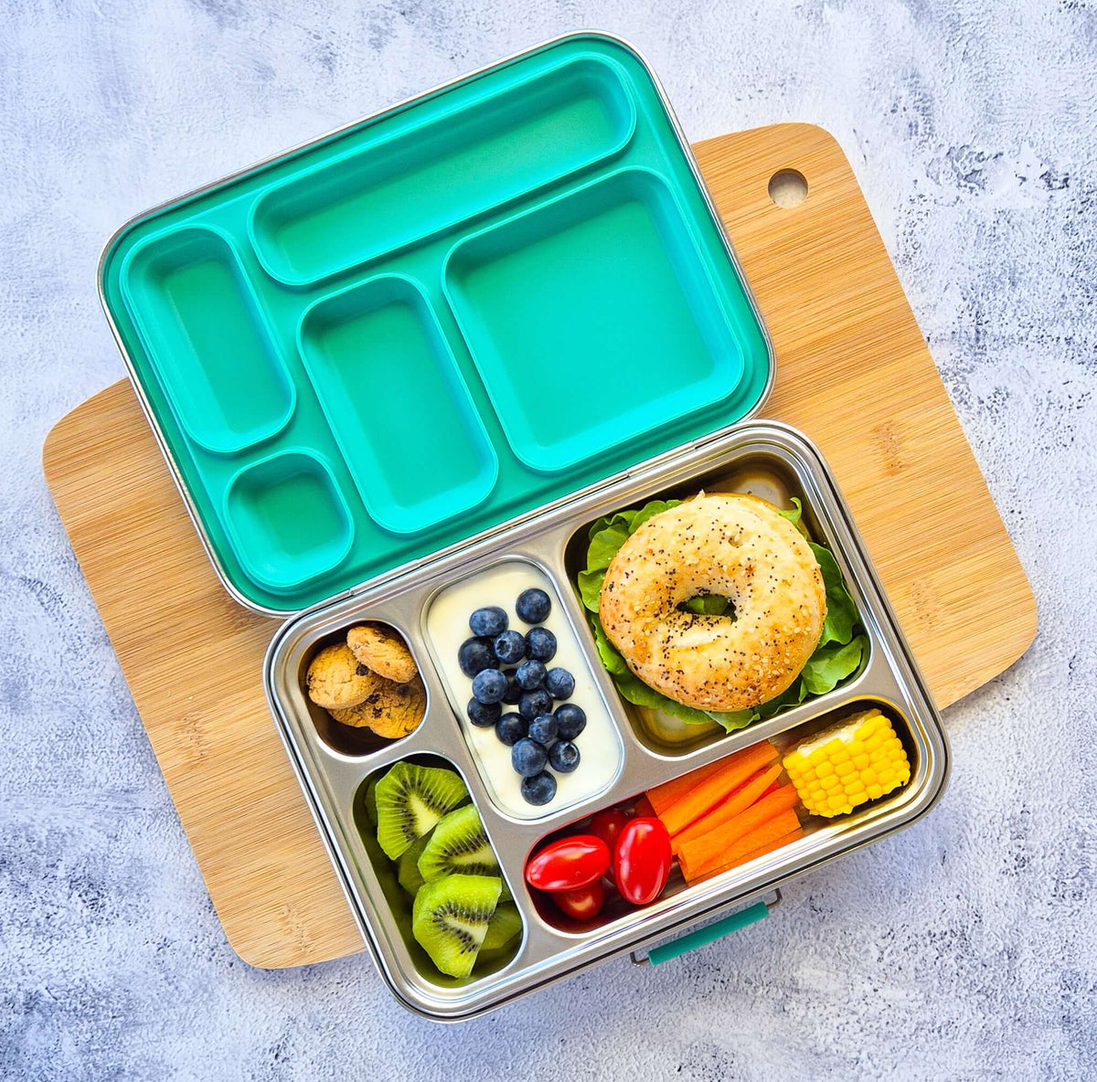 Five-compartment bento box with a teal lid on a wooden board, packed with a bagel, blueberries, kiwi, carrot sticks and corn