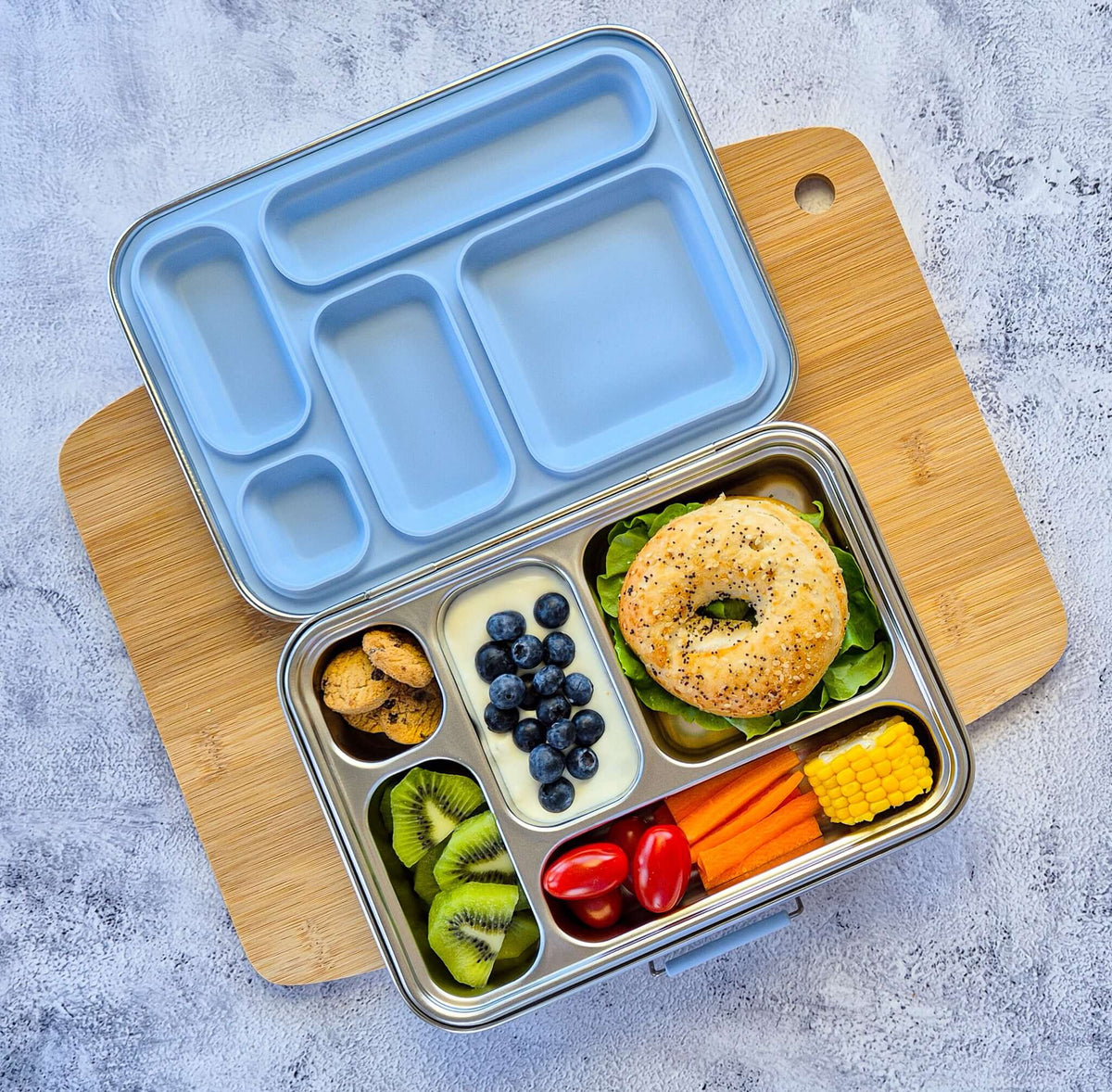 Five-compartment bento box with a pale blue lid on a wooden board, packed with a bagel, blueberries, kiwi and carrot sticks
