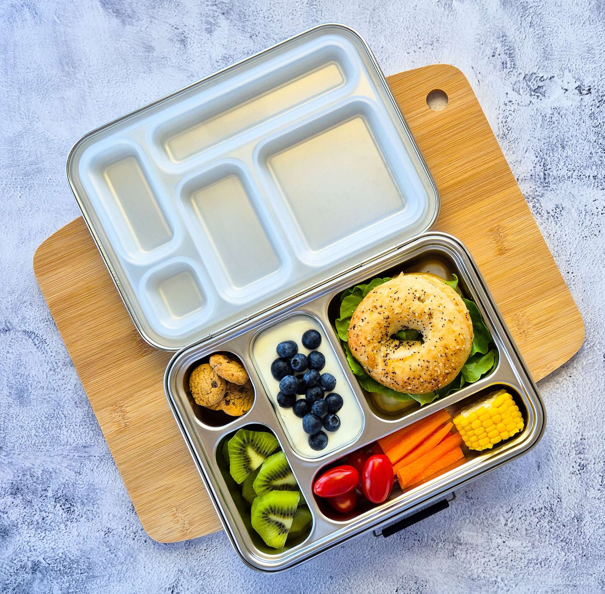 Five-compartment bento box with a pale blue lid on a wooden board, packed with a bagel, blueberries, kiwi and corn