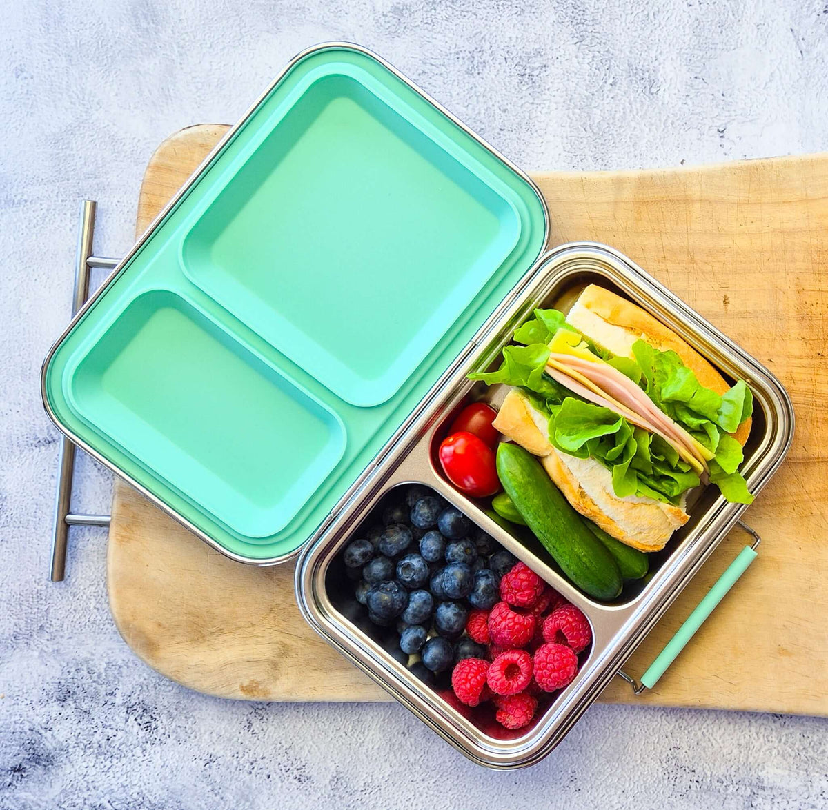 Two-compartment bento box with a mint lid on a wooden board, packed with a sandwich and berries