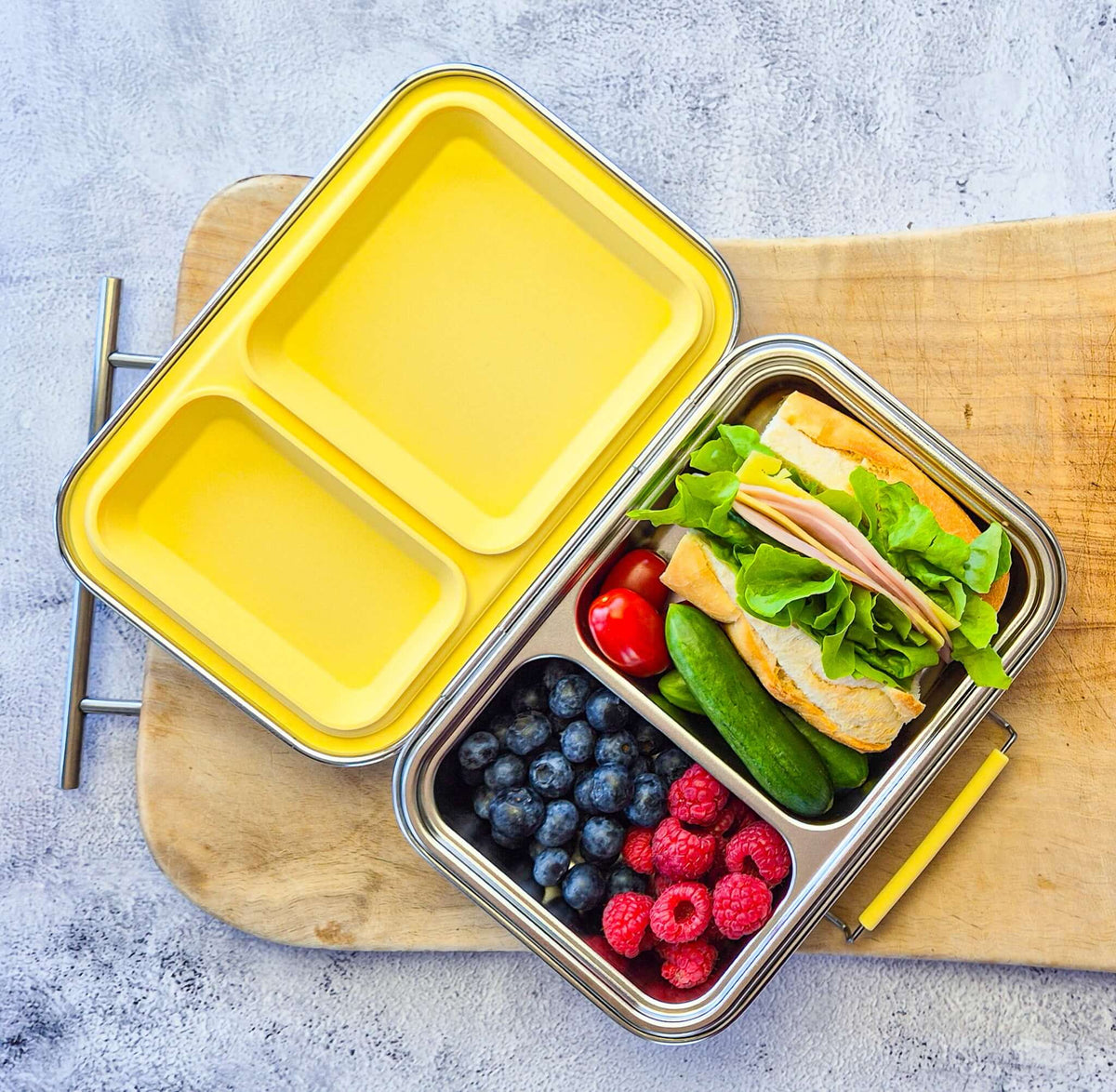 Two-compartment bento box with a yellow lid on a wooden board, packed with a sandwich, berries and cucumber
