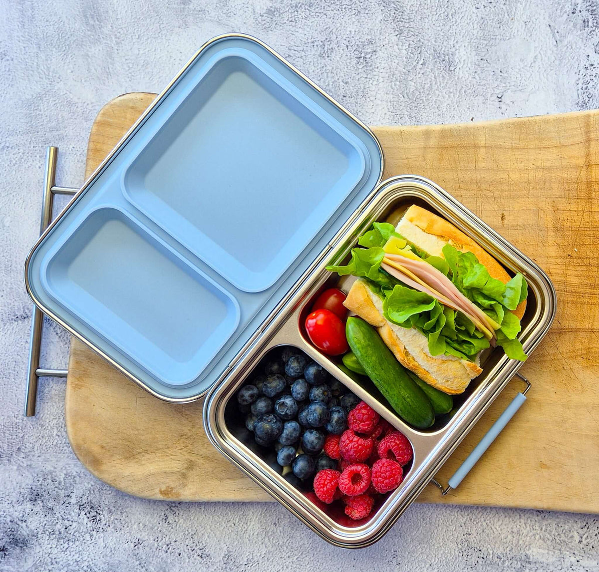 Two-compartment bento box with a pale blue lid on a wooden board, packed with a sandwich and berries