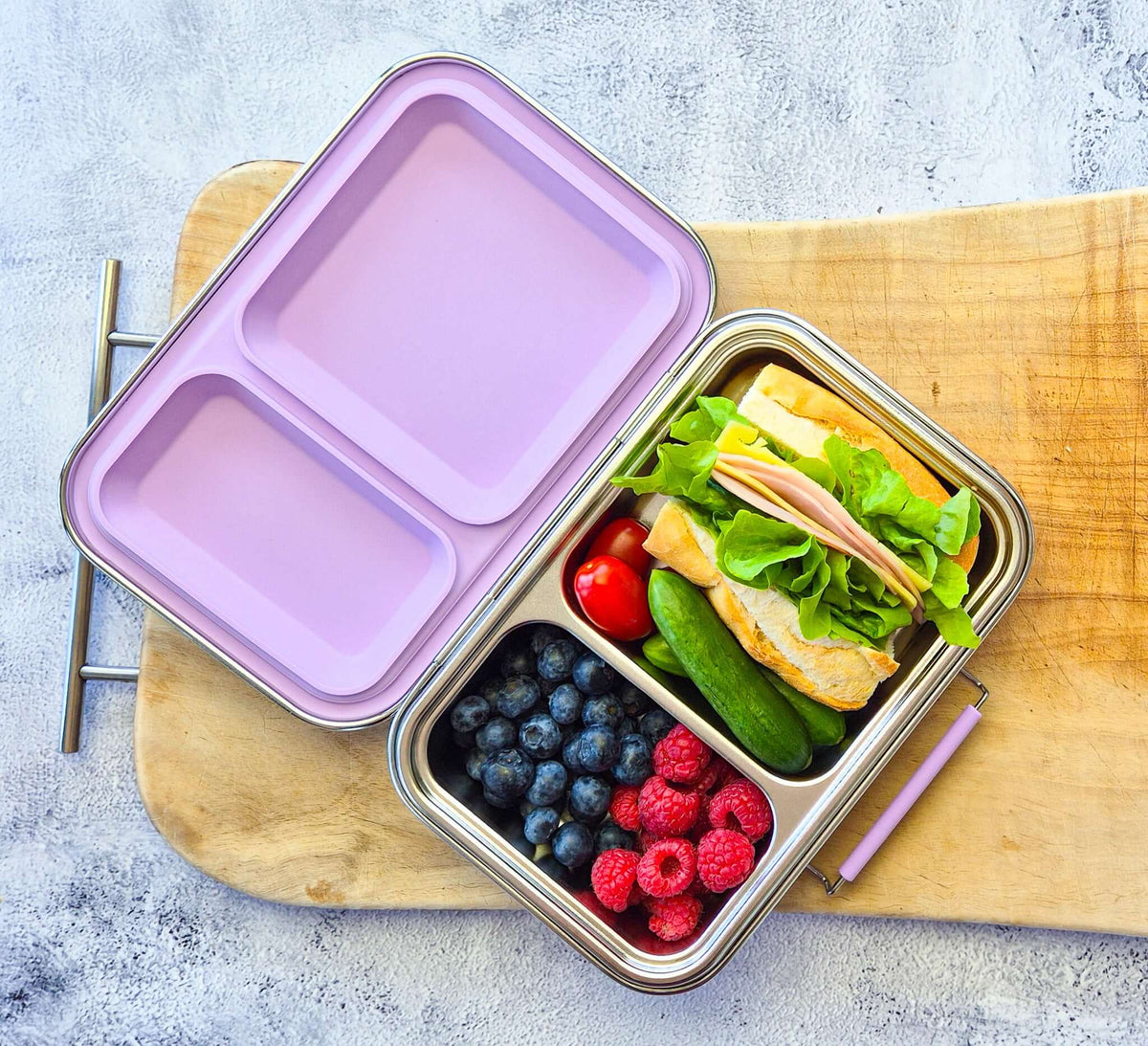 Two-compartment bento box with a lilac lid on a wooden board, packed with a sandwich and berries