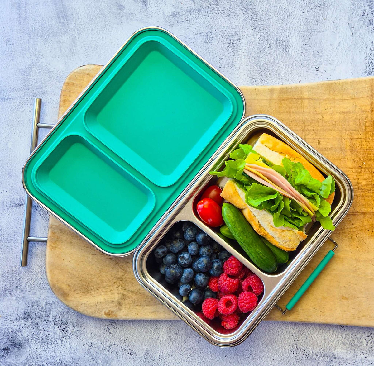 Two-compartment bento box with a teal lid on a wooden board, packed with a sandwich and berries