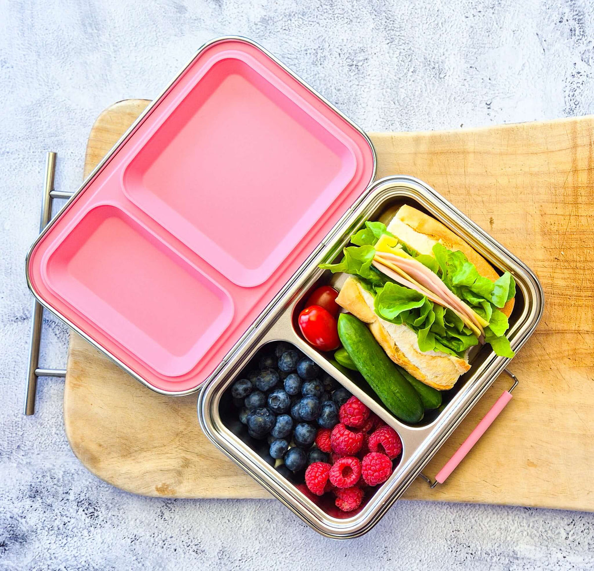 Two-compartment bento box with a pink lid on a wooden board, packed with a sandwich and berries