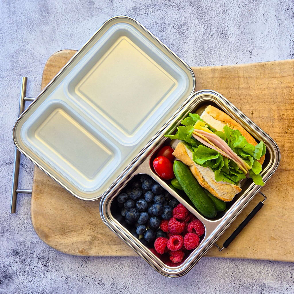 Two-compartment bento box with a clear lid on a wooden board, packed with a sandwich and berries