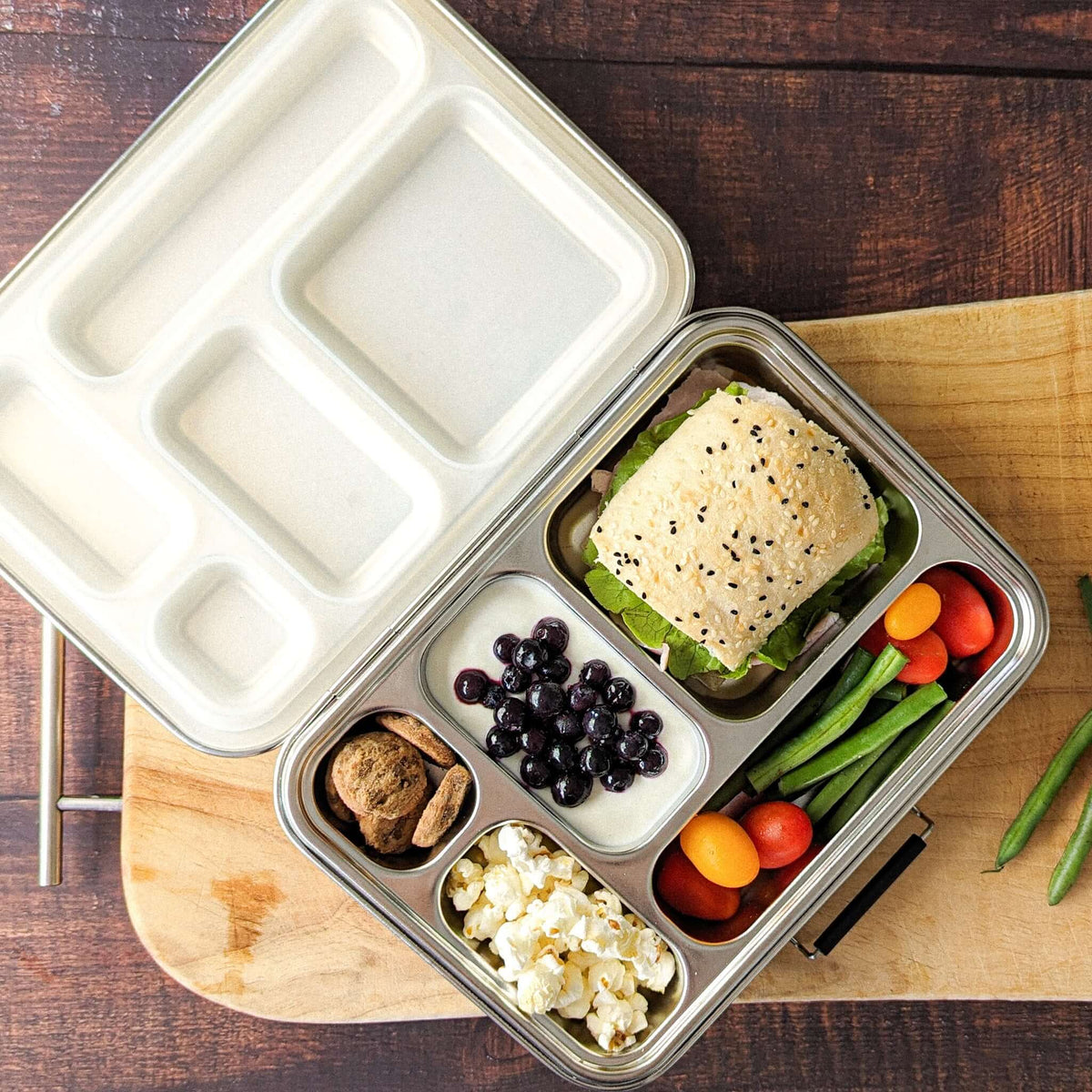 Five-compartment bento box with a cream lid packed with a sandwich, blueberries, popcorn, tomatoes and green beans