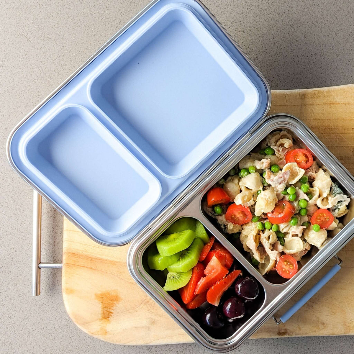 Two-compartment bento box with a pale blue lid packed with pasta salad, capsicum and olives
