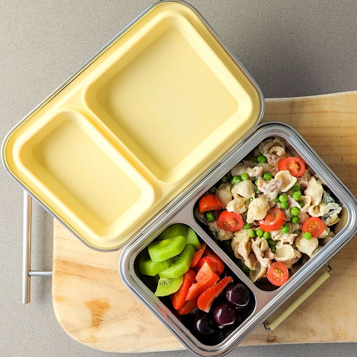 Two-compartment bento box with a yellow lid packed with pasta salad, capsicum and olives