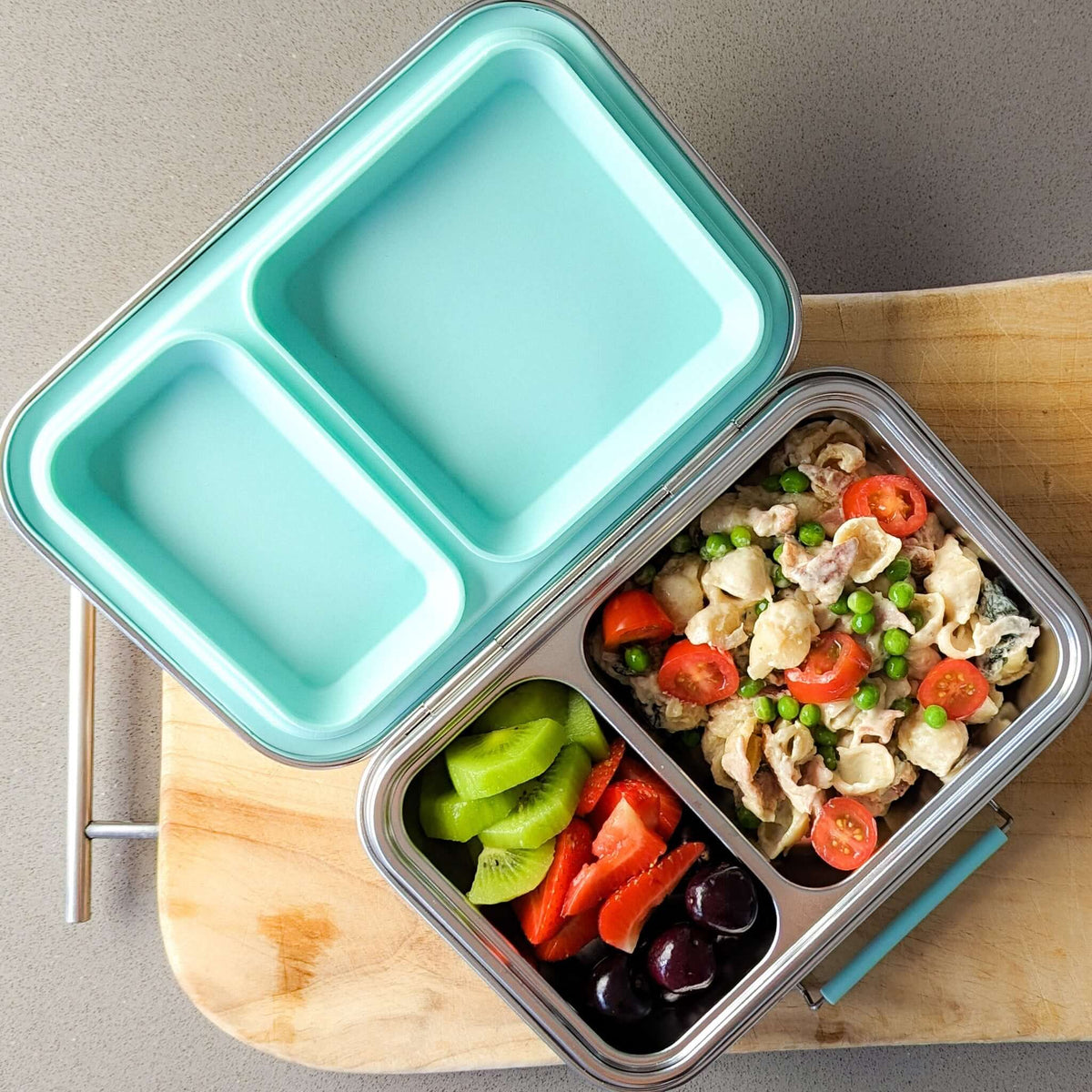Two-compartment bento box with an aqua lid packed with pasta salad, capsicum and olives