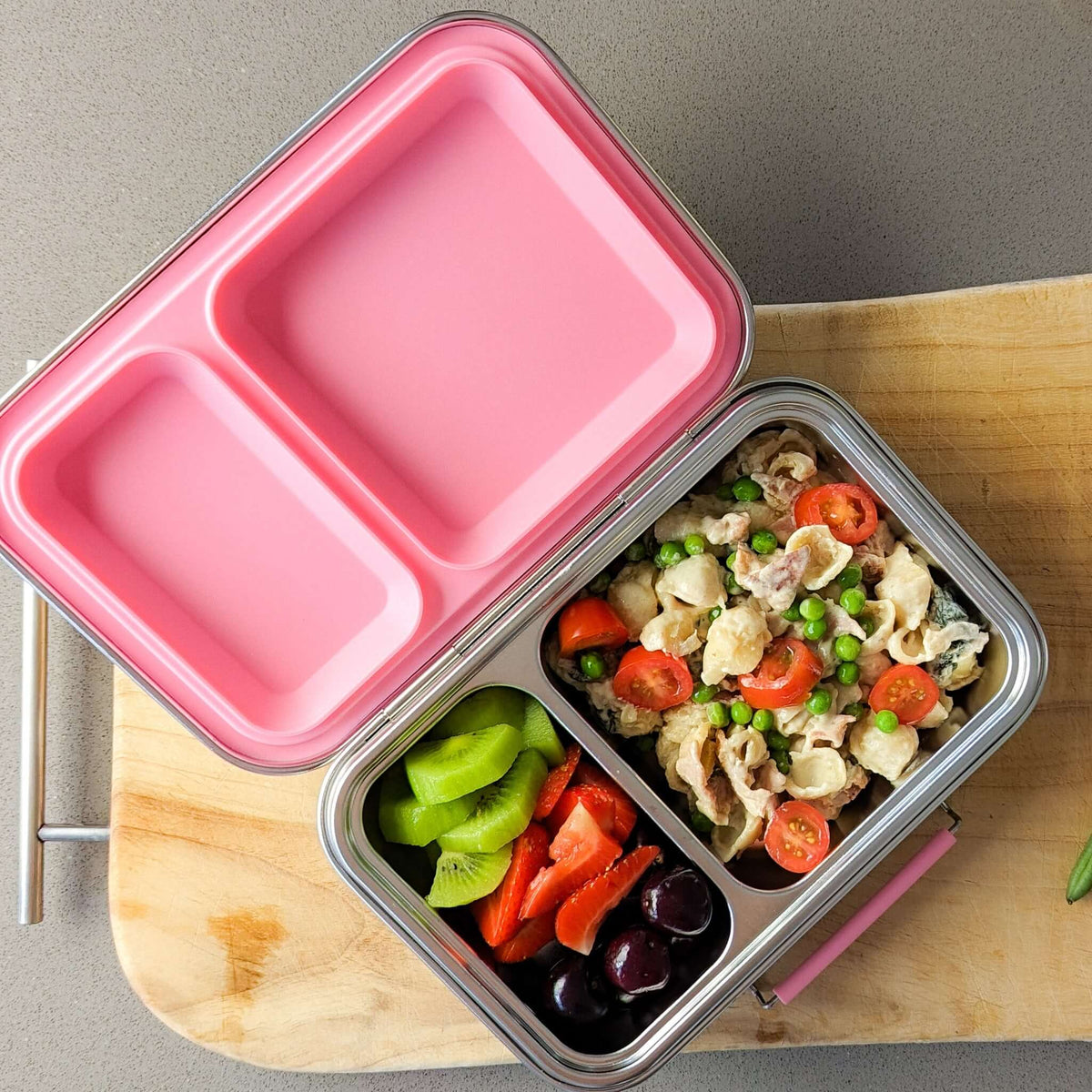 Two-compartment bento box with a pink lid on a wooden board, packed with pasta salad, capsicum and olives