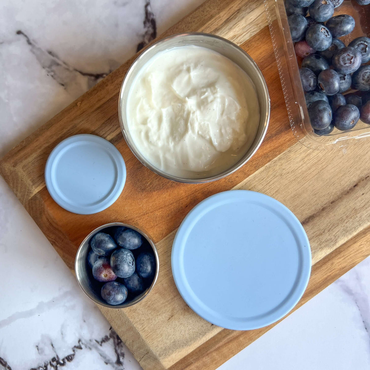 Two stainless steel snack pots with pale blue lids on a wooden board beside yoghurt and blueberries