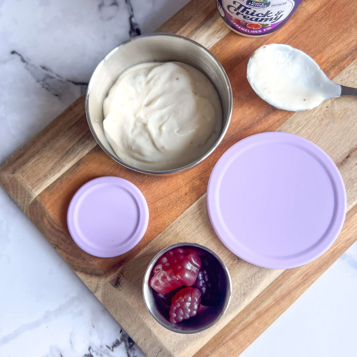 Stainless steel snack pots with lilac lids on a wooden board beside yoghurt and berries