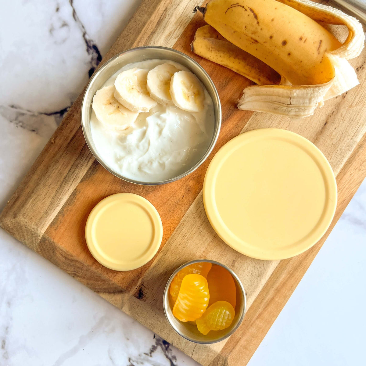 Stainless steel snack pots with yellow lids on a wooden board beside banana and custard