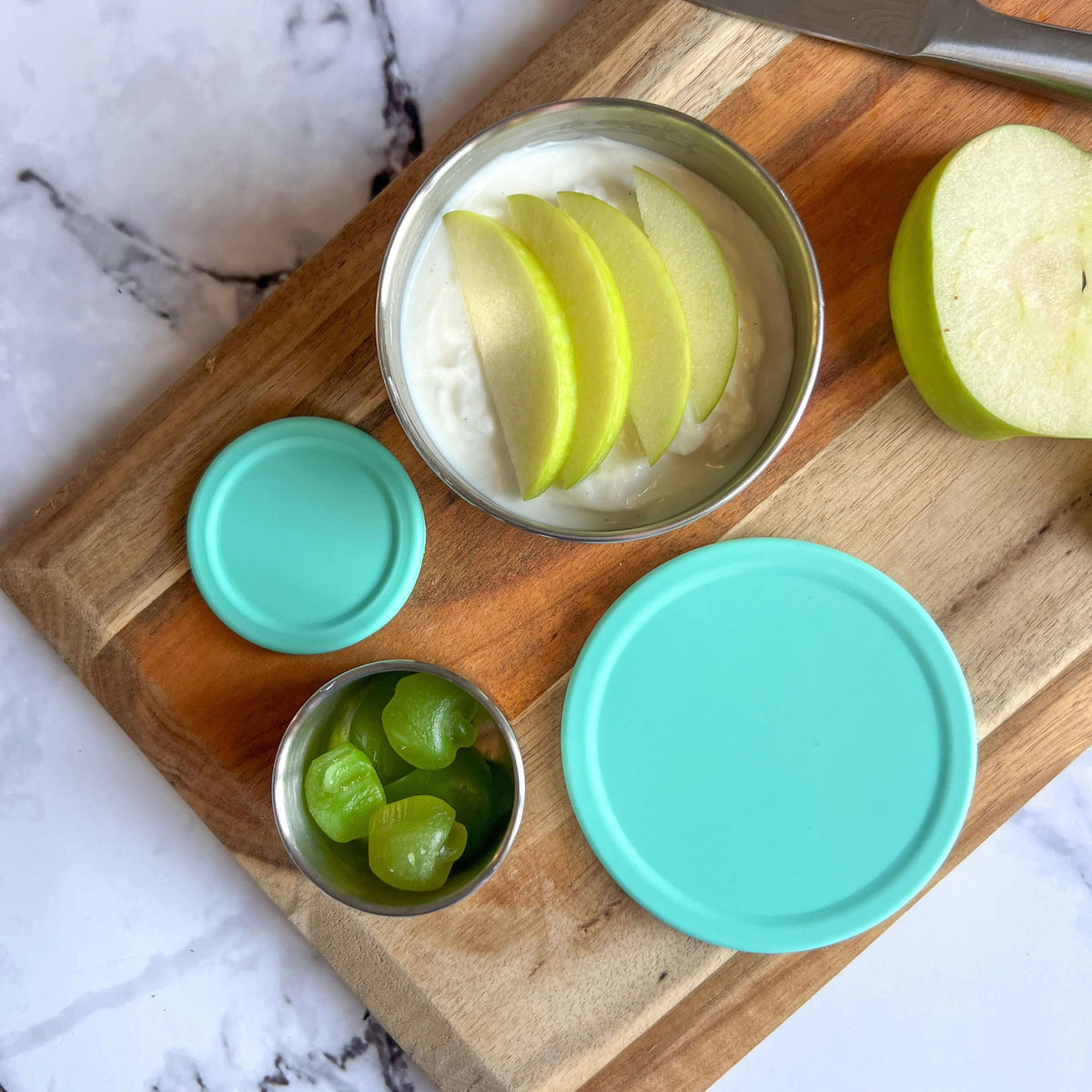 Stainless steel snack pots with turquoise lids on a wooden board beside apple slices and grapes