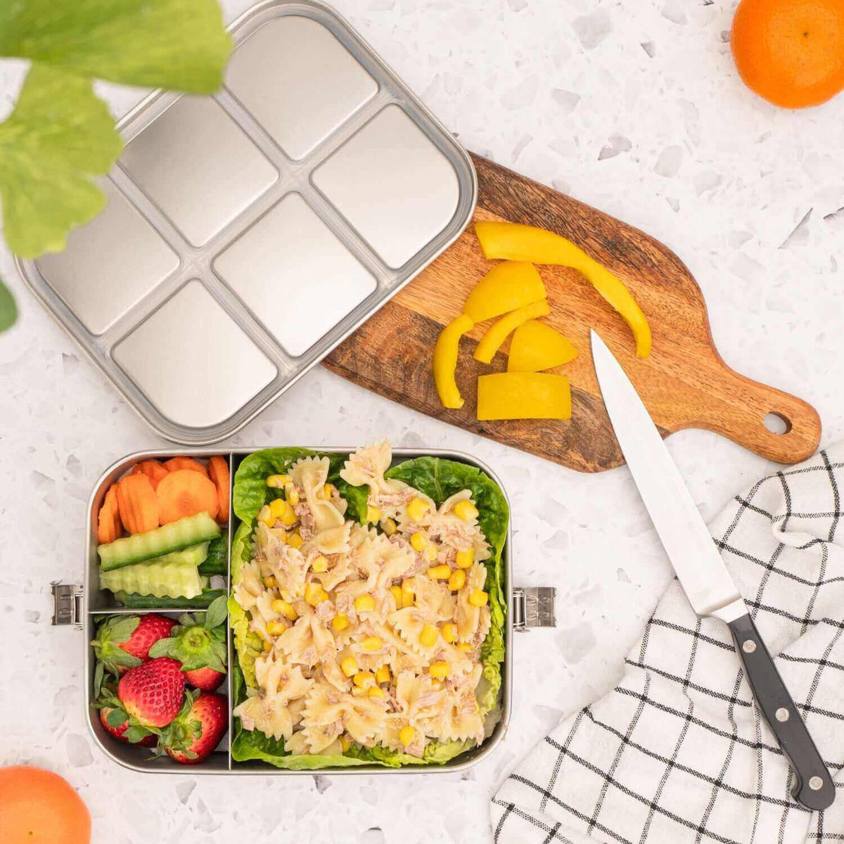 Stainless steel lunch box with compartments filled with salad and fruits, cutting board with sliced yellow bell pepper, and a knife on a white surface.
