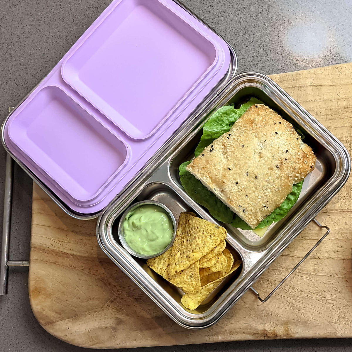 Two-compartment bento box with a lilac lid packed with a sandwich, corn chips and guacamole