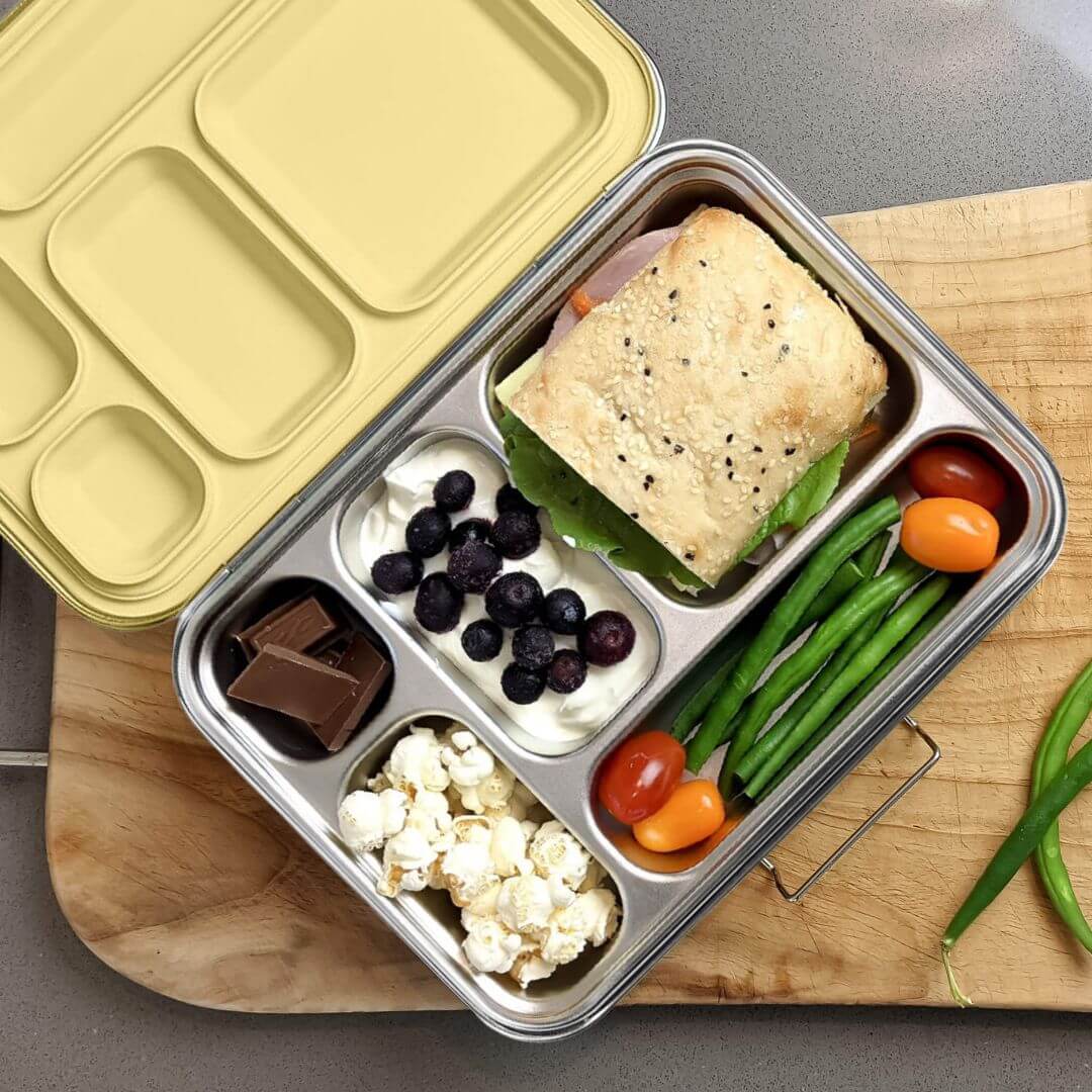 Five-compartment bento box with a yellow lid packed with a sandwich, blueberries, popcorn, tomatoes and green beans