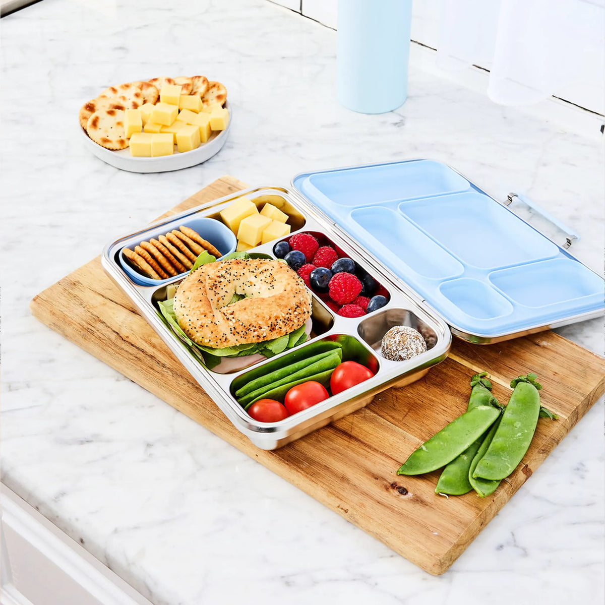 Bento lunch box with a pale blue lid on a wooden board, packed with a bagel, berries, tomatoes and snap peas