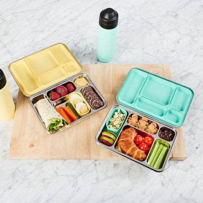 Two bento lunch boxes with yellow and mint lids on a wooden board, packed with fruit, vegetables and crackers