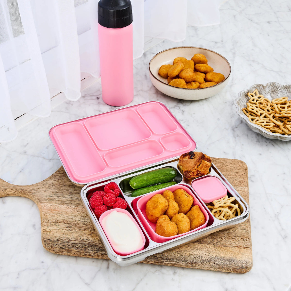 Bento lunch box with a pink lid on a wooden board, packed with chicken nuggets, cucumber and raspberries beside a pink drink bottle
