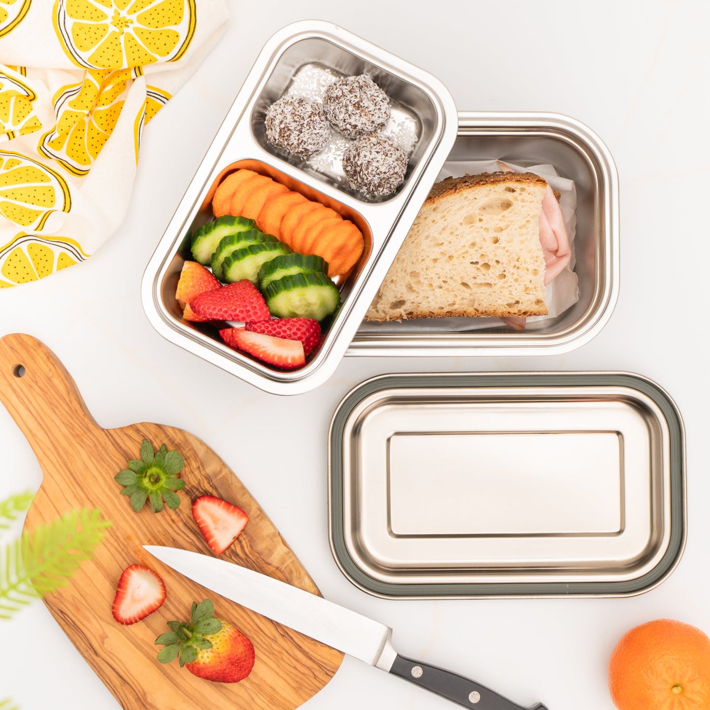 Metal lunch stainless steel container with compartments of food on a white surface with a wooden cutting board and knife.