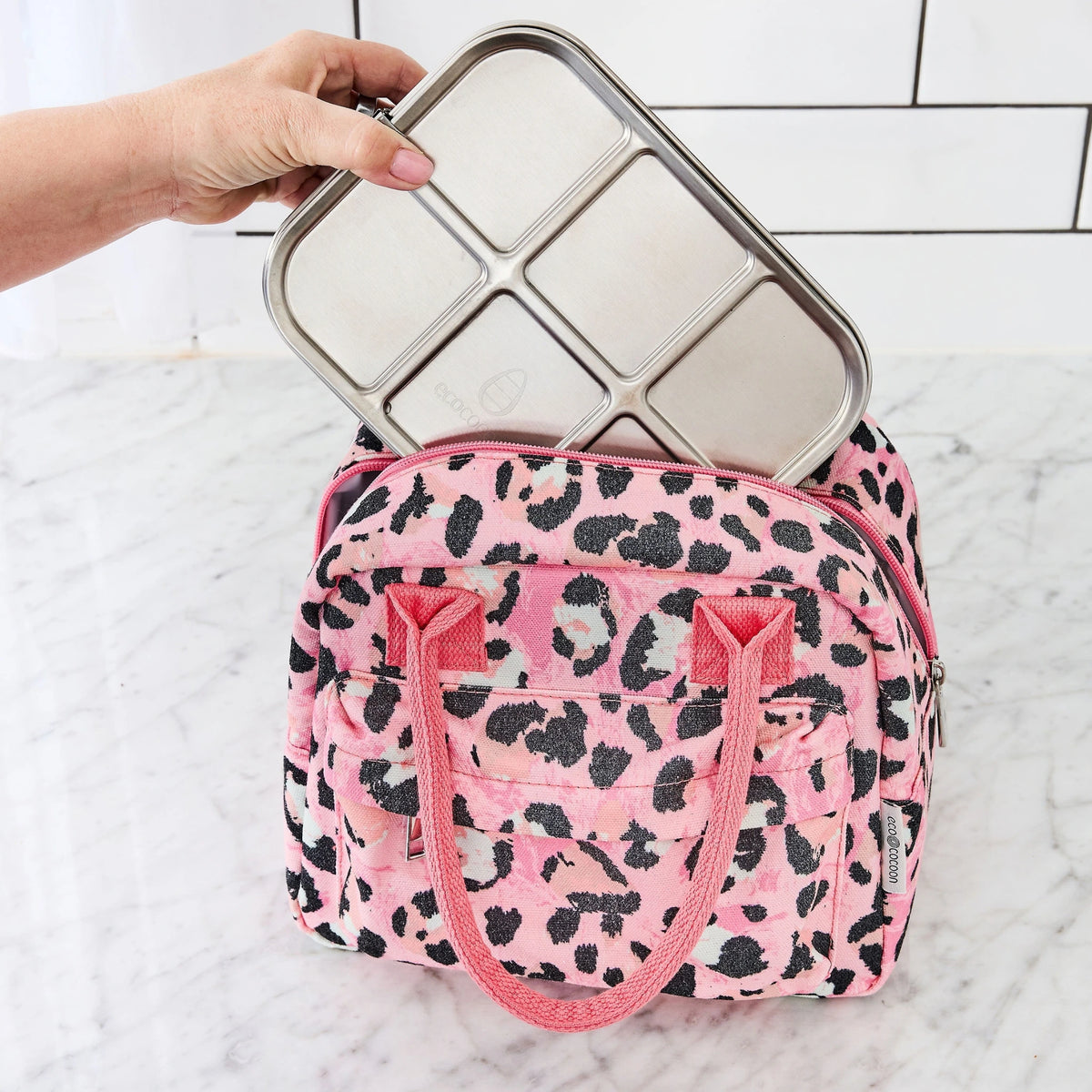Hands lowering a stainless steel bento box into a pink leopard print lunch tote