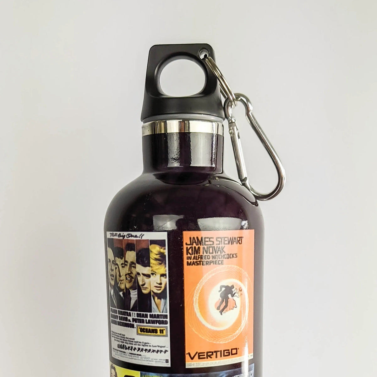 A dark purple insulated water bottle with a black lid and carabiner, featuring a James Stewart Kim Novak film poster label on the front, set against a plain light gray background.