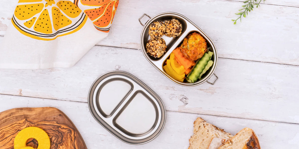 An oval stainless steel divided lunch box with two handles, shown open on a white wood surface with food compartments containing salad, granola, fruit, and sandwich bread. The lid is placed beside it, and a napkin and decorative board are partially visible.