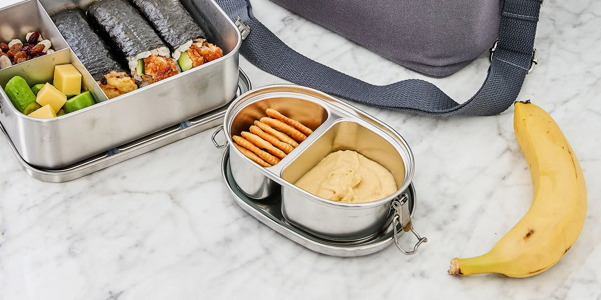 Two stainless steel compartment lunch boxes on a white marble surface, one filled with sushi, vegetables, crackers, and dip, and the other open with crackers and dip. A gray bag and a banana are partially visible around them.