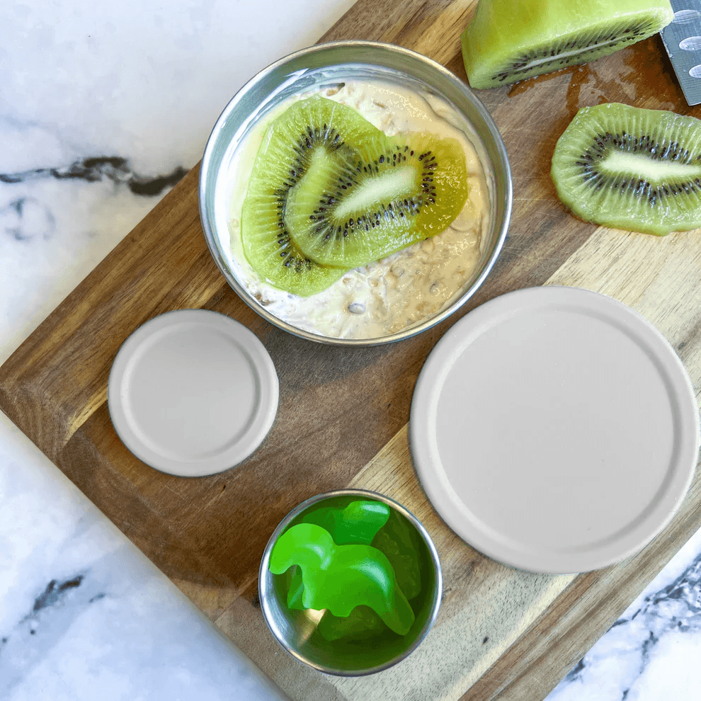 A stainless steel lunch box set arranged on a wooden cutting board over a white marble surface. The set includes a main compartment with oatmeal topped with kiwi slices, a small cup with bright green gelatin or candy, and two beige silicone lids.