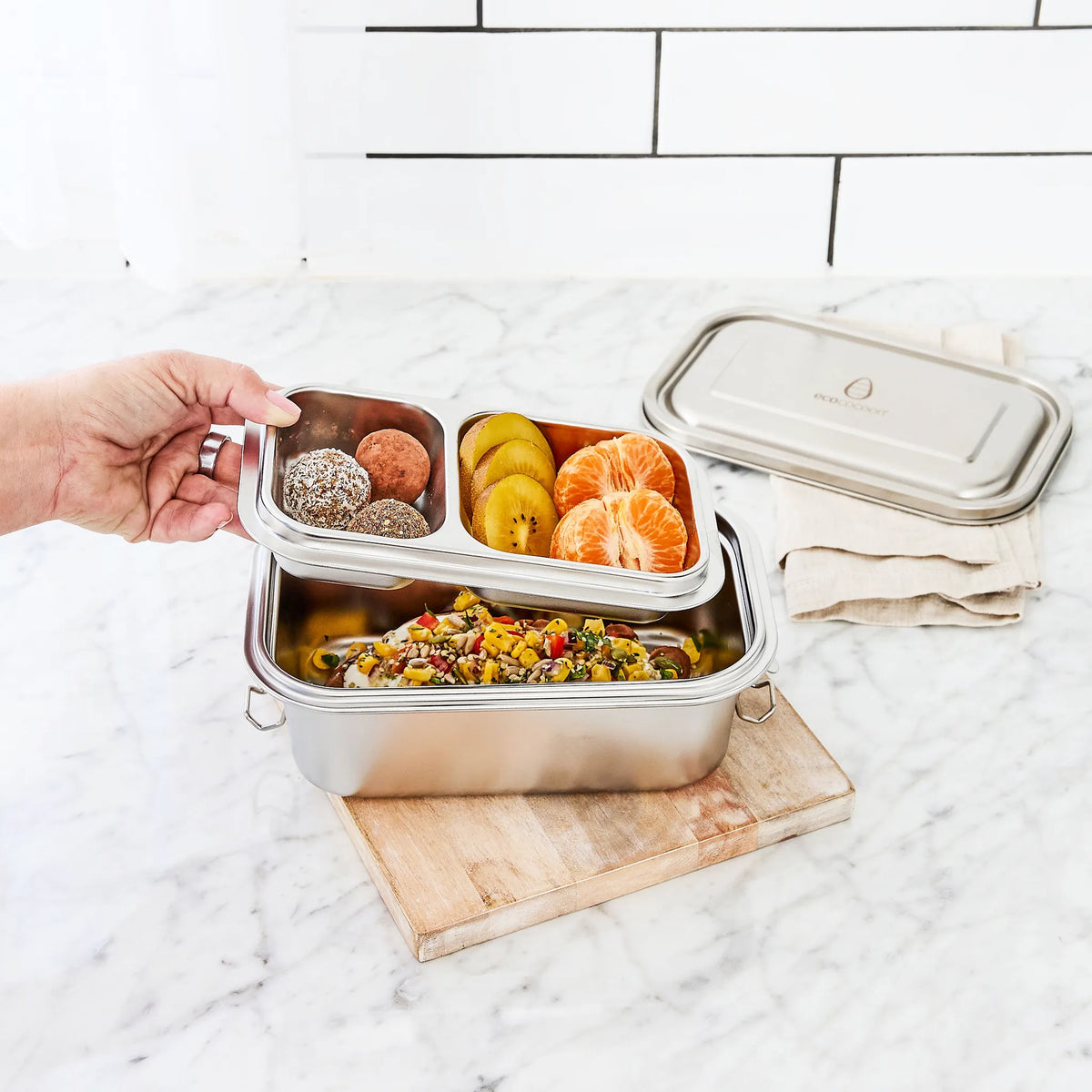 Stainless steel bento lunch box on a wooden board on a marble benchtop, with a hand lifting out a removable two-compartment tray. The matching lid rests on a folded linen napkin nearby.