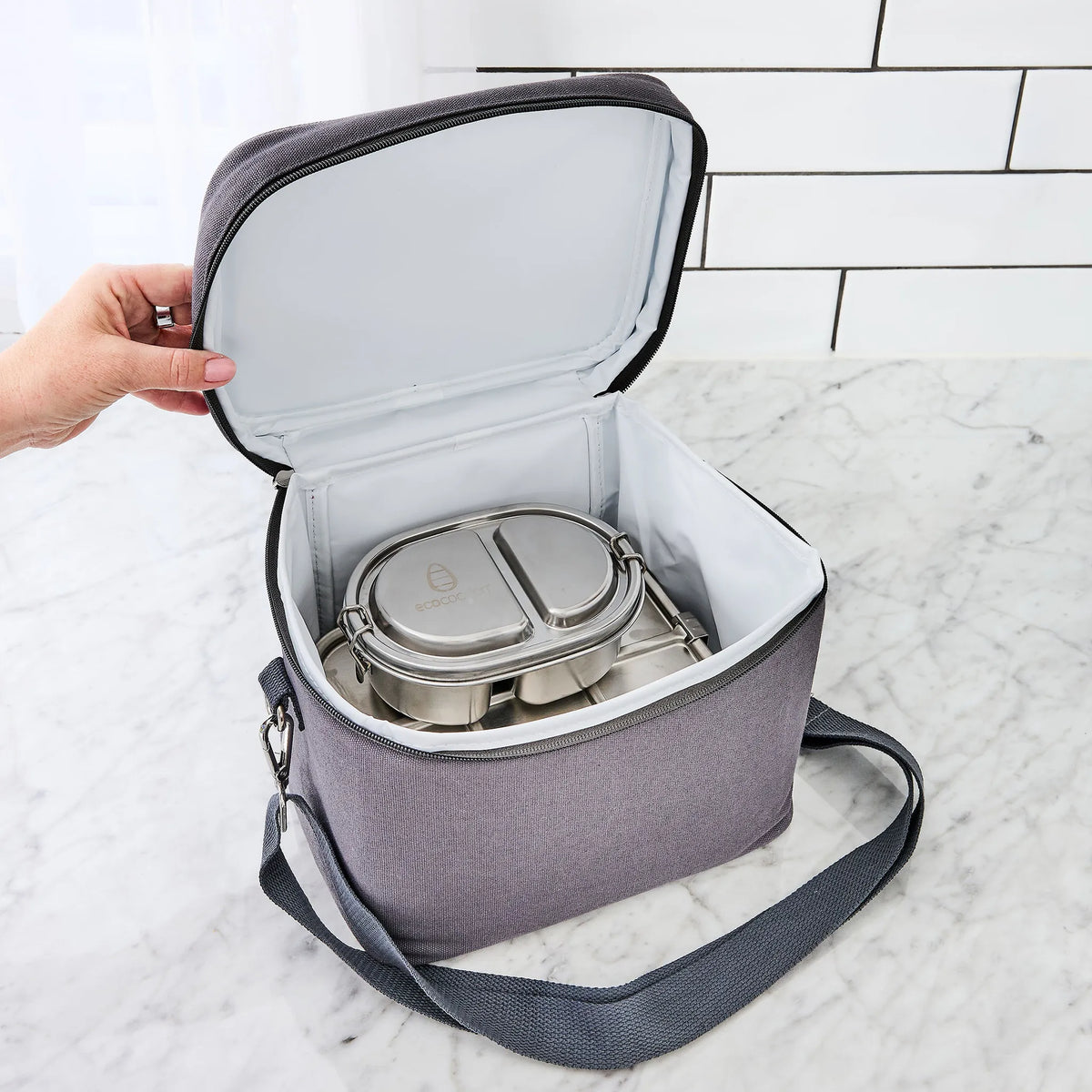 A gray insulated lunch bag with a white interior, shown open with a stainless steel bento container inside. A hand is holding the left side of the bag, and the bag is placed on a white marble surface with a white subway tile wall in the background.