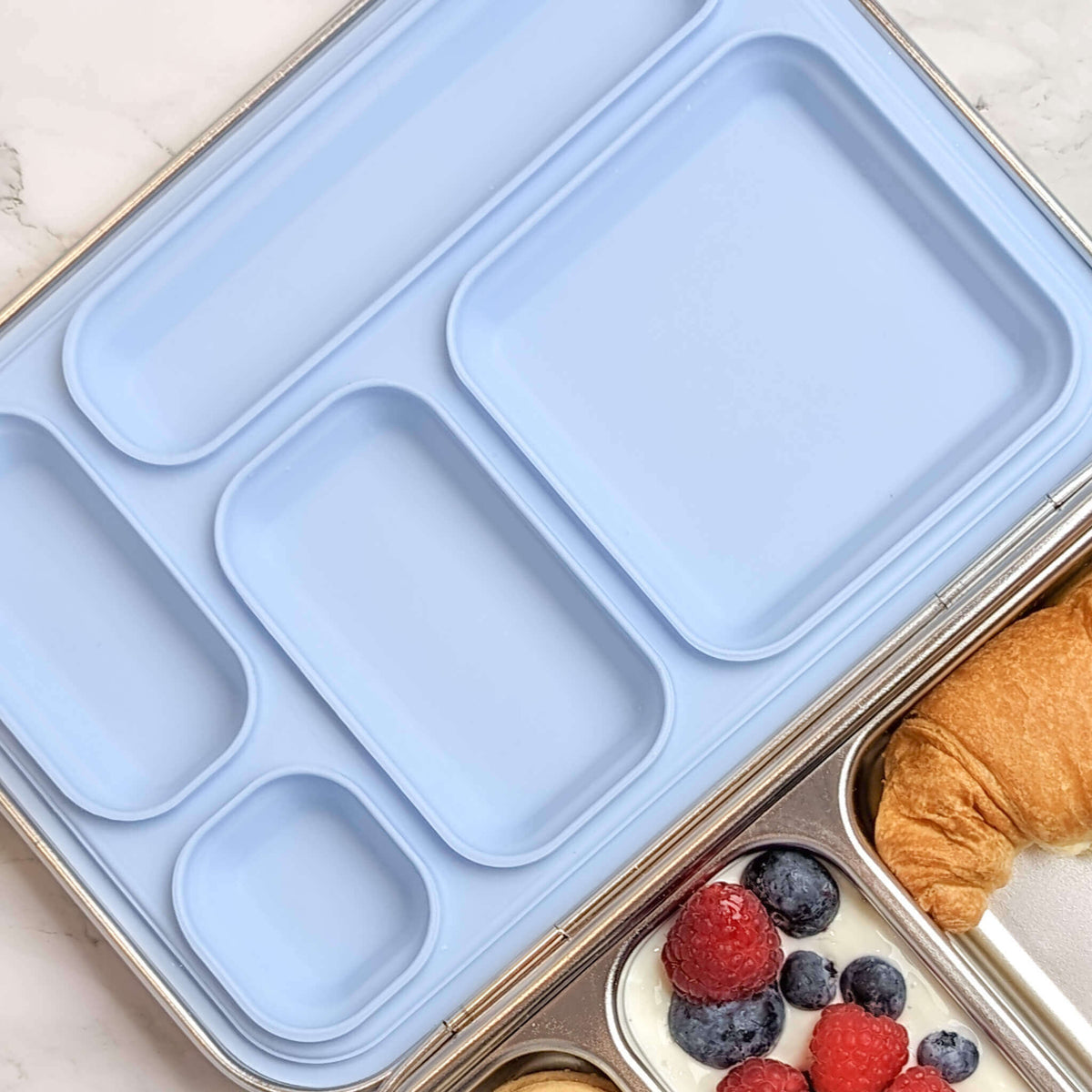 Pale blue replacement silicone seal in a five-compartment bento lid beside berries and a croissant