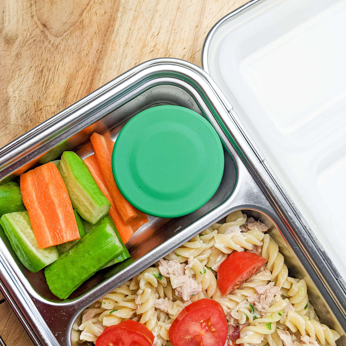 Green-lidded snack pot sitting inside a bento box beside pasta salad, carrot and celery sticks