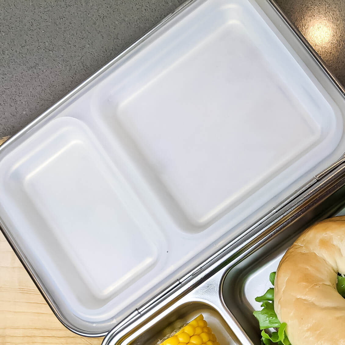 White replacement silicone seal in a two-compartment bento lid beside a packed sandwich