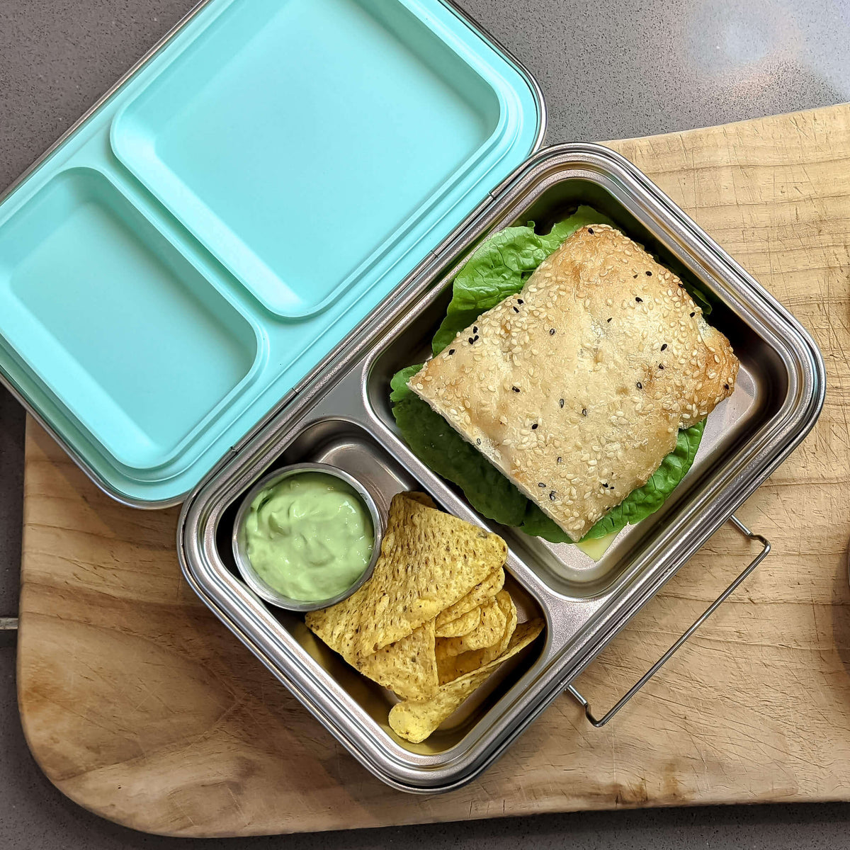 Two-compartment bento box with a pale blue lid on a wooden board, packed with a sandwich, corn chips and guacamole