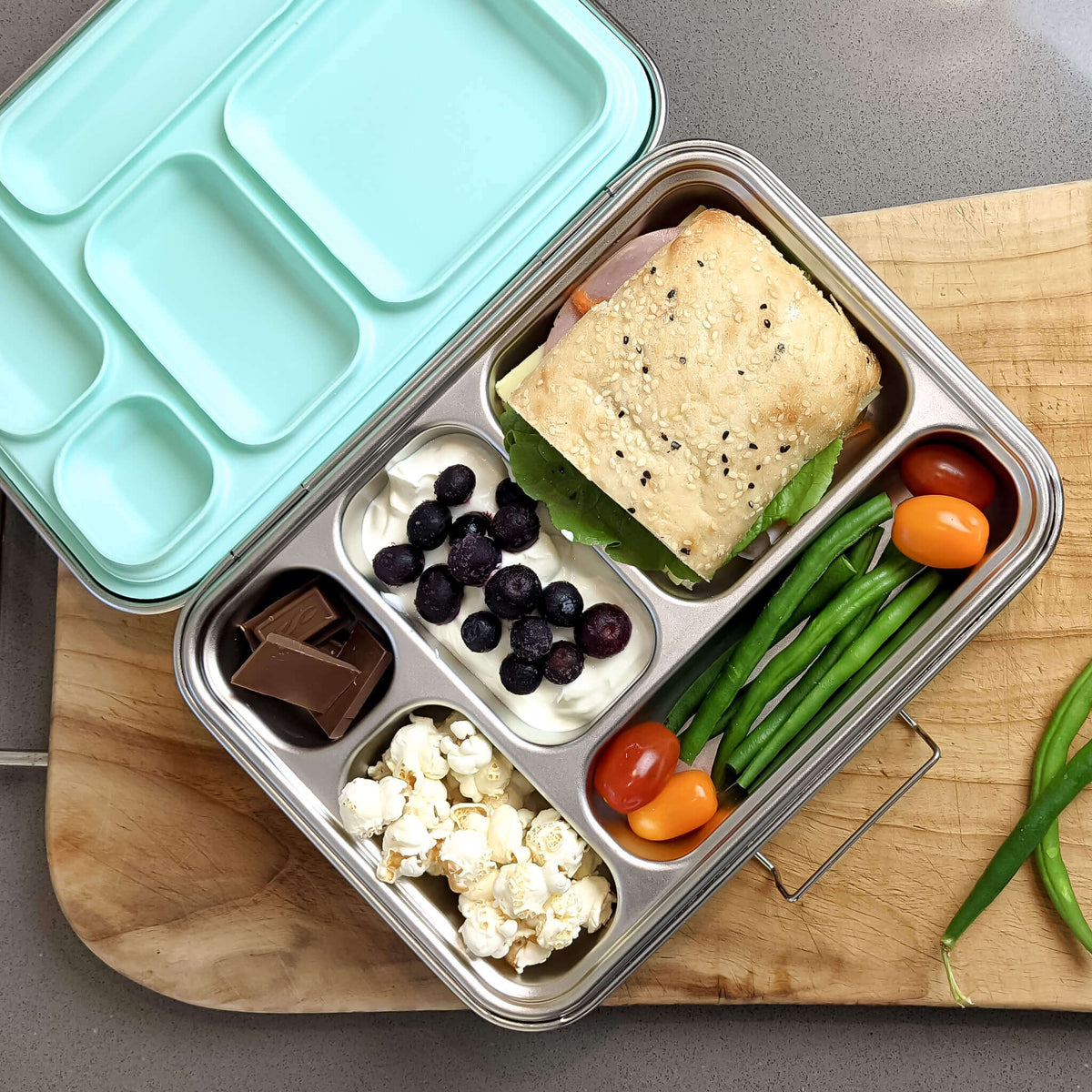 Five-compartment bento box with a pale blue lid packed with a sandwich, blueberries, popcorn, tomatoes and green beans